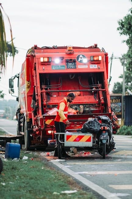 A worker wearing a yellow high-visibility vest and dark trousers is standing behind a red rubbish collection truck on a roadside during the daytime. The truck, with identifiable safety features such a