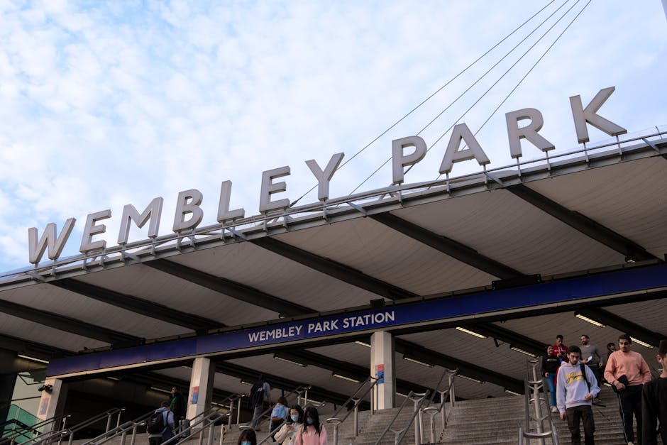 The image displays the exterior of Wembley Park Station, with large, bold, uppercase metal letters spelling 'WEMBLEY PARK' mounted on the roof above the entrance steps. Below the sign, there is a blue